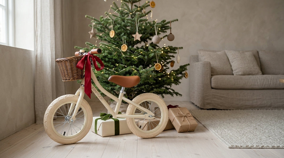 Cream Banwood balance bike with a red bow beside a Christmas tree and wrapped presents
