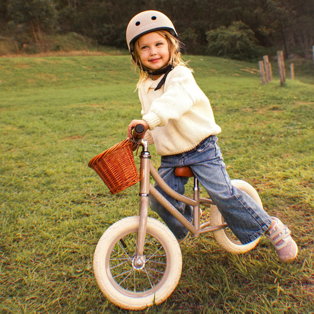 Child in cream jumper and helmet riding beige balance bike with red basket on grass