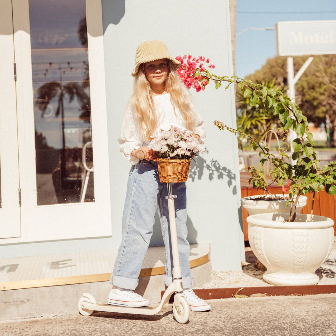 Child riding cream Banwood scooter with wicker basket in sunny outdoor coastal setting