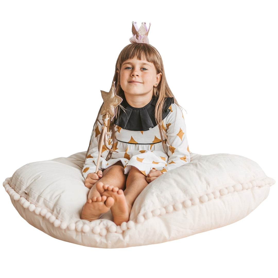 A girl in costume sitting on the MINICAMP ecru pom pom floor cushion