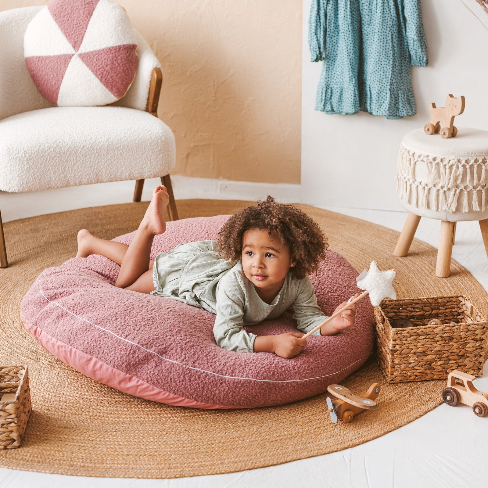 Child lying on the MINICAMP rose bouclé floor cushion in a boho-style nursery with a cream bouclé armchair and wicker baskets
