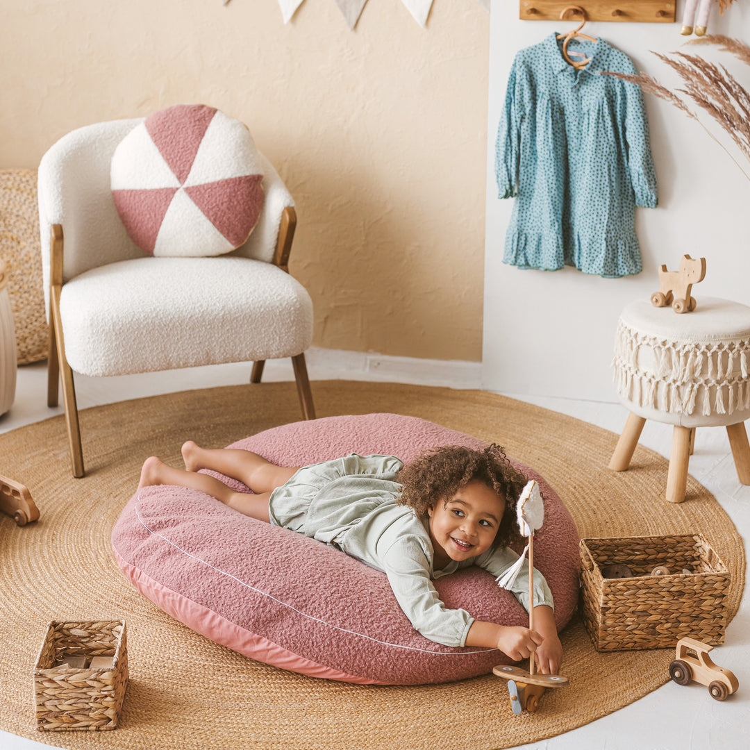 Child lying face down on the MINICAMP rose bouclé floor cushion, smiling, in a nursery playroom setting
