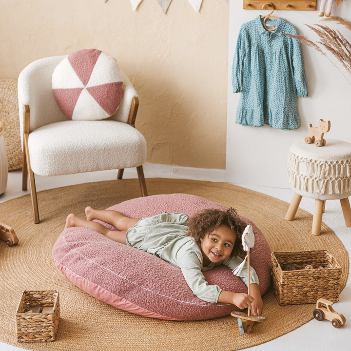 Child lying face down on the MINICAMP rose bouclé floor cushion, smiling, in a nursery playroom setting