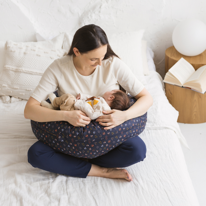 Woman sitting on a bed nursing her baby supported by the MINICAMP Navy Blue floral C-Shape Pillow