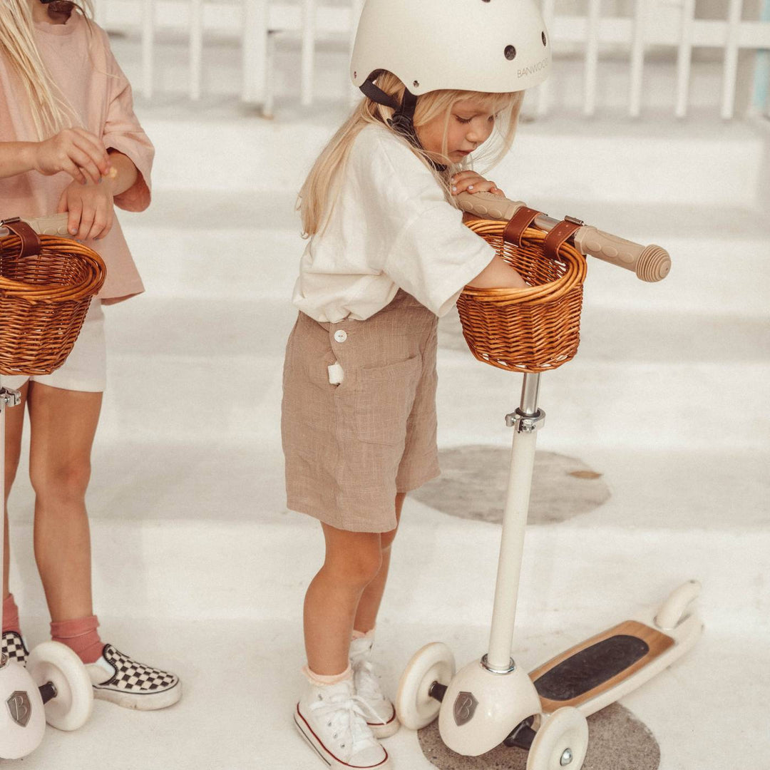 Two children riding cream Banwood scooters with wicker baskets in outdoor play setting