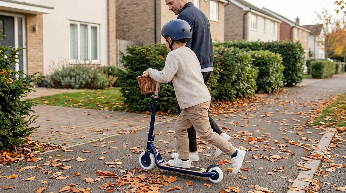 Child in a helmet riding a navy Banwood Maxi Scooter Vintage along an autumn pavement with an adult alongside